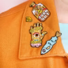 Una chaqueta naranja adornada con broches de esmalte de colores, entre ellos un pez, una mano con texto y una botella.