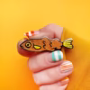 Una mano sujeta un alfiler de esmalte en forma de pez con detalles de purpurina naranja.