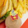 Pin de esmalte con forma de pez dentro de una caja de patatas fritas, presentado en una tarjeta con una hamburguesa y patatas fritas de fondo.