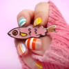 Una mano con un jersey rosa sujetando un alfiler de esmalte con forma de pez de purpurina rosa.