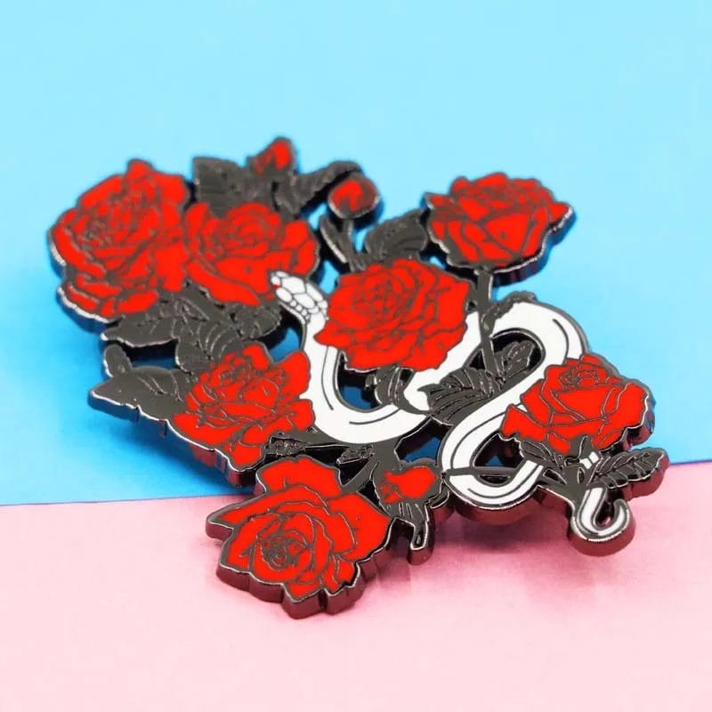 Alfiler de esmalte de rosa roja con una serpiente entrelazada alrededor de un corazón, que simboliza el amor y la transformación.