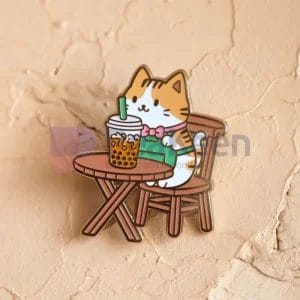 Un pin's mignon représentant un chat assis à une table avec une tasse de thé, conçu comme un pin's personnalisé de chat boba.