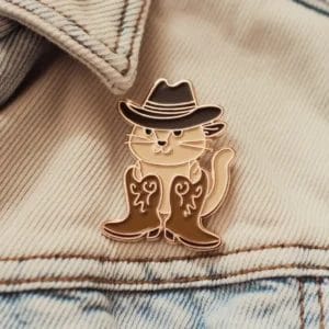 Una spilla con un gatto vestito con stivali da cowboy e cappello, etichettata come "Cowboy Cat Pins".