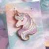 Spilla unicorno visualizzata su un cartoncino con un vivace sfondo arcobaleno.